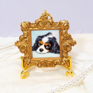 Puede incluir: Un pequeño retrato cuadrado de un perro Cavalier King Charles Spaniel tricolor dentro de un marco dorado ornamentado. El marco presenta intrincados adornos y volutas, y se exhibe sobre un soporte de metal dorado a juego.