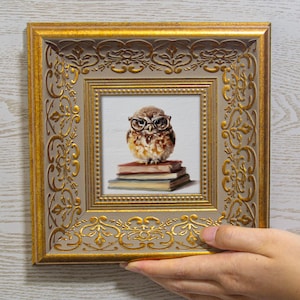 May include: A square wall art piece featuring a painting of a brown owl wearing round glasses perched on a stack of three books. The artwork is encased in a decorative gold frame with intricate embossed patterns and a light beige inner border.