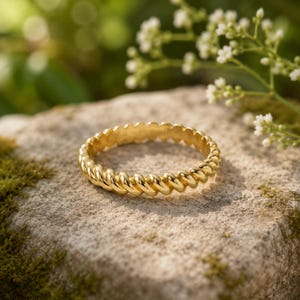 Op de afbeelding: Een gepolijste gouden ring met een doorlopend gedraaid touwontwerp. De band heeft een uniforme, afgeronde textuur over de gehele omtrek, waardoor een reeks onderling verbonden, schuine ribbels ontstaat die het licht op het oppervlak vangen.