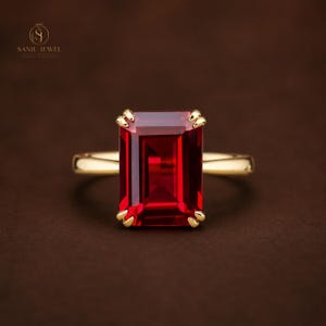 Puede incluir: Un anillo de oro con una gran piedra preciosa roja de corte esmeralda rectangular sujeta por cuatro garras. La banda es de oro liso y pulido. El texto SANJU JEWEL es visible en la esquina superior izquierda junto a un pequeño logotipo circular.