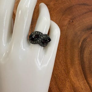 May include: A ring featuring a dark metal band with an infinity-style crossover design. The surface is encrusted with small, shimmering grey stones and accented by two prominent, deep red, oval-shaped faceted gemstones set within the center.