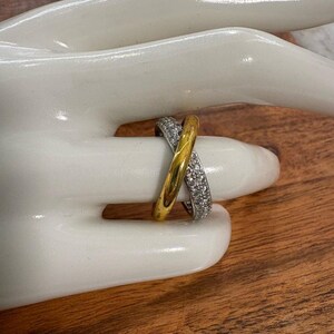 May include: A crossover ring featuring two bands, one smooth polished gold band and one silver-toned band encrusted with small clear stones, creating an interlocking design that wraps around the finger in a crisscross pattern.