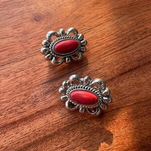 May include: A pair of silver-toned earrings featuring an oval-shaped red stone center. Each earring is surrounded by a decorative twisted rope border and a scalloped metal frame with openwork loops, creating a detailed and textured appearance.