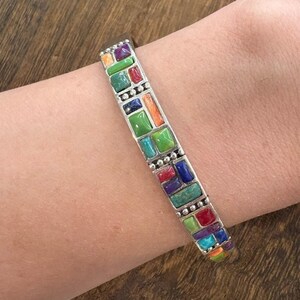 May include: A silver cuff bracelet featuring a mosaic inlay of rectangular and square stones in vibrant shades of green, blue, purple, red, and orange. The metal frame includes small silver bead accents between the colorful stone segments.