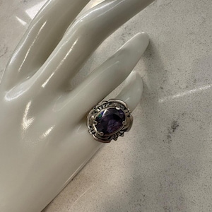 May include: A silver ring featuring an oval-shaped, deep purple faceted gemstone set in a decorative metal band with intricate cut-out details and prongs securing the stone in the center of the setting.
