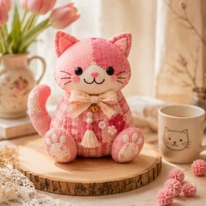 May include: A pink and white patchwork cat plush toy with a bow, sitting on a wooden slice. The cat has embroidered details and a bell. A matching mug with a cat face and pink flowers are also in the image.