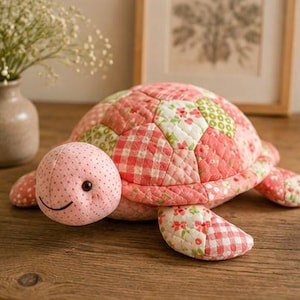 May include: A plush turtle toy featuring a quilted shell made of various pink, red, and white fabric patches with floral, gingham, and polka dot patterns. The head is light pink with small black eyes and a stitched smile, and the flippers match.