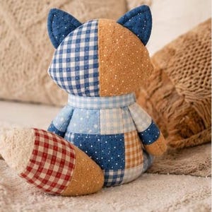 May include: A patchwork fox plush toy in various fabrics. The fox has blue and white gingham, dotted tan, and a red and white checked tail. The toy is on a soft, textured surface. A charming handmade item.