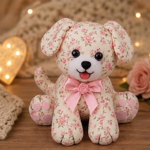 May include: A plush dog toy made of cream-colored fabric with a pink floral pattern. The toy features large black plastic eyes, a white muzzle, a black nose, and a pink tongue. A pink satin ribbon with a small fabric rose is tied around its neck.