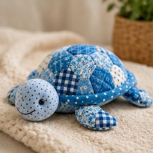Peut inclure: Une tortue en peluche avec une carapace en patchwork composée de divers tissus à motifs bleus et blancs, incluant des carreaux vichy, des fleurs et des pois. La tête est bleu clair avec de petits pois noirs, un sourire brodé et des yeux en perles.