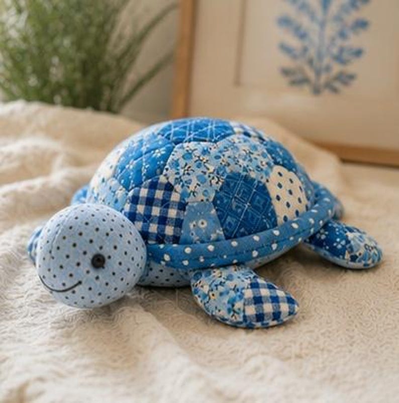 May include: A stuffed turtle toy crafted from various blue and white patterned fabrics. The shell features a quilted patchwork design with floral, gingham, and polka dot prints. The head is light blue with small dark polka dots and a black button eye.