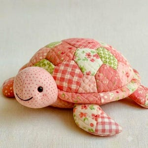May include: A stuffed turtle toy crafted from quilted fabric patches in shades of pink, cream, and light green. The shell features a patchwork design of floral and gingham patterns, while the head is light pink with small black dot eyes.