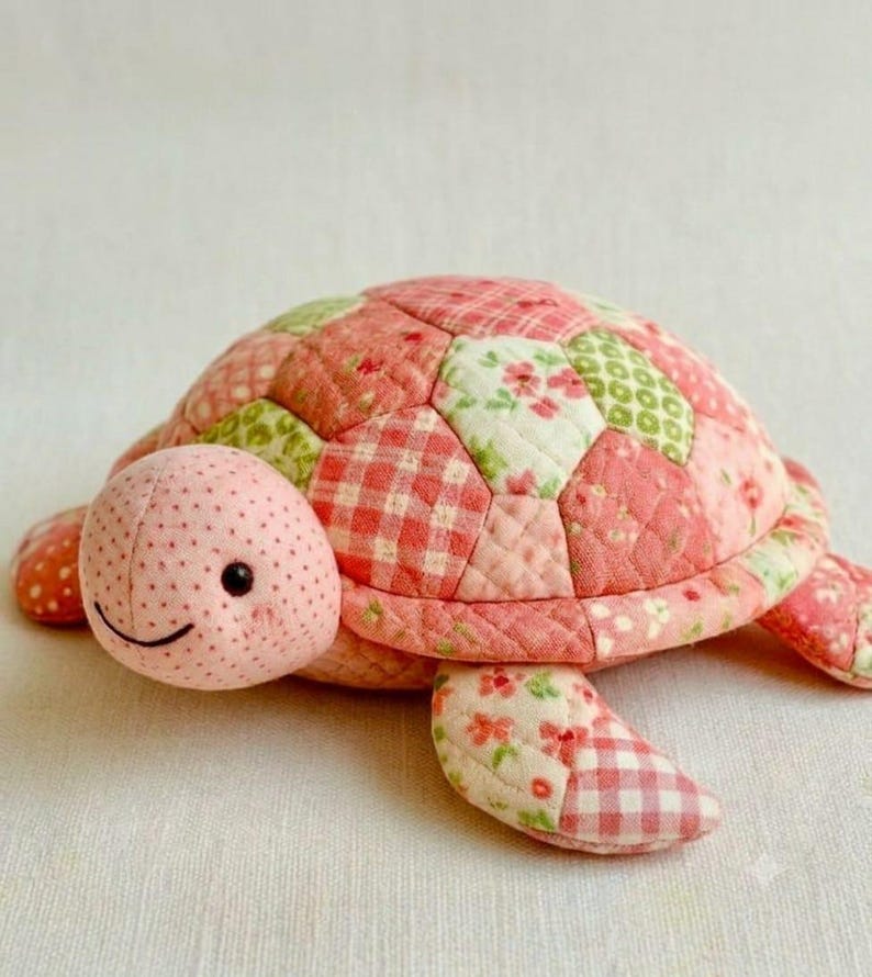May include: A stuffed turtle toy crafted from quilted fabric patches in shades of pink, cream, and light green. The shell features a patchwork design of floral and gingham patterns, while the head is light pink with small black dot eyes.