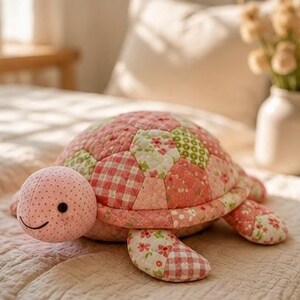 May include: A plush turtle toy featuring a quilted patchwork shell in shades of pink, white, and green with floral and gingham patterns. The head is light pink with small dark polka dots, a simple stitched black smile, and round black eyes.