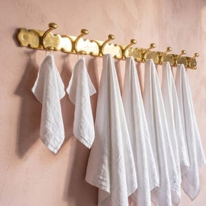 May include: A long, horizontal brass wall rack featuring seven curved hooks. Each hook holds a single white cloth. The brass rack has a scalloped edge design and is mounted to a wall. The seven white cloths hang vertically from the brass hooks.