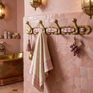 May include: A wall-mounted brass coat rack featuring four hooks and decorative rectangular inlays with pink and white geometric patterns. A beige towel with a pink patterned border hangs from one hook, alongside a small tassel and dried flowers.
