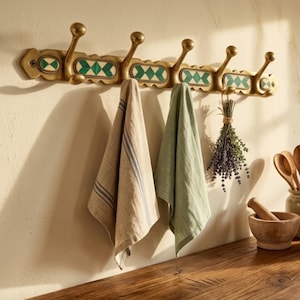 Peut inclure: Un porte-manteau mural en laiton doté de cinq crochets et d'incrustations rectangulaires décoratives à motif géométrique vert et blanc. Une serviette beige rayée, une serviette vert sauge et un bouquet de lavande séchée sont suspendus.