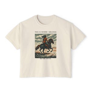 Könnte beinhalten: Cremefarbenes T-Shirt mit einem Aufdruck einer Person, die auf einem schwarzen Pferd reitet. Der Aufdruck enthält den Text "THIS IS WHERE I BELONG" und "IN THE WIND". Das Shirt zeigt eine Person, die durch ein Feld reitet. Das Shirt ist aus weichem Material.