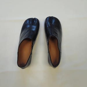 May include: A pair of black leather shoes featuring a distinct split-toe design at the front. The shoes have a smooth, dark exterior finish and a tan-colored interior lining, showcasing a low-profile silhouette with a rounded toe shape.