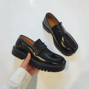 Pode incluir: Um par de mocassins de couro preto brilhante com um design distinto de biqueira dividida, uma tira tradicional de mocassim sobre o peito do pé e uma sola grossa e tratorada com ranhuras profundas para maior altura e construção robusta.
