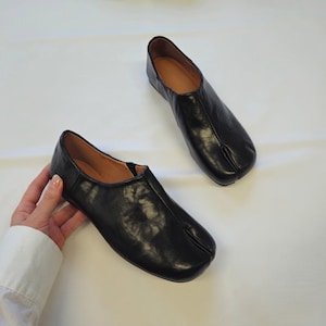 Könnte beinhalten: Ein Paar schwarze Slipper mit einem markanten Zehensteg-Design. Die Schuhe haben eine glatte, dunkle Oberfläche mit einer sichtbaren Naht, die mittig über den Vorderfuß bis zur Zehentrennung verläuft. Das Innenfutter ist in einem Beigeton gehalten.