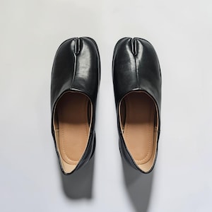 Puede incluir: Un par de zapatos de cuero negro con un diseño distintivo de punta dividida en la parte delantera. Los zapatos tienen un acabado exterior liso y pulido, una silueta redondeada y un forro interior de color tostado claro.