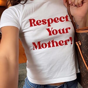 Peut inclure: Haut court blanc avec le texte rouge "Respect Your Mother!". Le t-shirt est associé à un jean bleu. Un sac à main marron avec un motif monogramme est visible sur l'image.