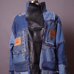以下が含まれることがあります： 黒い革のインサートが付いたパッチワークデニムジャケット。ジャケットは、さまざまな色合いの青いデニムパッチ、ぼろぼろの縁、茶色の革のLevi'sブランドタグが特徴です。ジャケットはマネキンに展示されています。