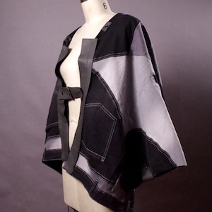 May include: A patchwork denim jacket in black and gray, displayed on a mannequin. The jacket features a unique design with contrasting panels, visible stitching, and a belt closure. The garment has a modern, artistic aesthetic.