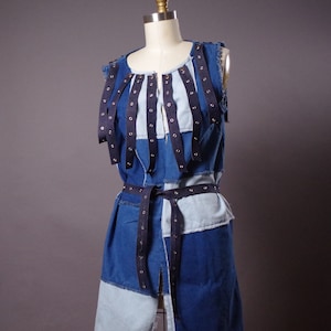 May include: A sleeveless denim dress with a patchwork design in various shades of blue. Dark blue straps with silver grommets are attached to the bodice. A matching belt cinches the waist. The dress is displayed on a mannequin.