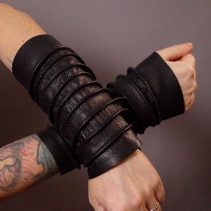 May include: A pair of black leather arm warmers with a pleated design. The arm warmers are worn on both arms and are crossed over each other.