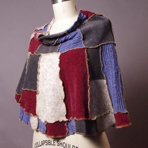 May include: A patchwork poncho in shades of gray, red, and blue, displayed on a mannequin. The poncho features a cowl neck and a mix of textures, including knit and felted fabrics. The design has a unique, handcrafted appearance.