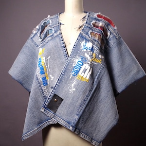 May include: A light blue denim jacket with a unique, deconstructed design. The jacket features distressed patches, paint splatters, and text graphics. A black leather patch is sewn on the front. The jacket is displayed on a mannequin.