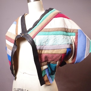 May include: A patchwork bolero jacket with a variety of colorful fabric strips. The jacket features a black leather strap closure and is displayed on a mannequin. The color palette includes red, blue, green, and tan.