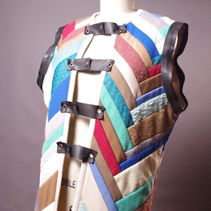 May include: A colorful, patchwork vest with a variety of fabric textures and colors, including blue, red, and brown. The vest features black leather straps with metal rivets and a black trim along the shoulders. The word "SIBLE" is visible on the vest.