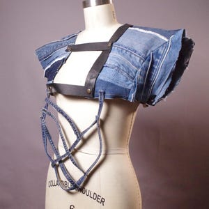 May include: A denim top with a unique design. The top is constructed from various shades of blue denim, featuring a black leather harness and intricate denim detailing. The garment is displayed on a mannequin.