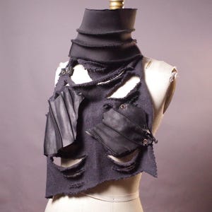 May include: A dark gray, distressed neck warmer with a high, layered collar. The garment features cut-out sections and is accented with black leather patches and metal rivets. The item is displayed on a mannequin.