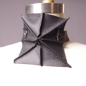 May include: A black leather collar with a geometric design. The collar is made of multiple triangular pieces that meet in the center. It has a strap with eyelets for closure and is displayed on a mannequin.