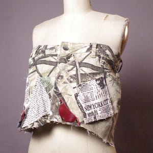 May include: A unique, handmade top constructed from repurposed camouflage fabric and denim. The garment features patches of newspaper print and red leather accents. The top is displayed on a mannequin, showcasing its distinctive design and construction.