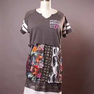 May include: A gray t-shirt with white stripes on the sleeves and the text "Philadelphia Phillies" and a red "P" on the front. The t-shirt is layered over a patchwork skirt with a variety of colorful patterns, including eyes, flowers, and geometric designs.
