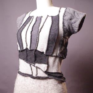 以下が含まれることがあります： グレーのパッチワークトップス、半袖付き。明るいグレーと濃いグレーの生地が幾何学模様に縫い合わされています。袖はケーブル編みで、マネキンに着せています。