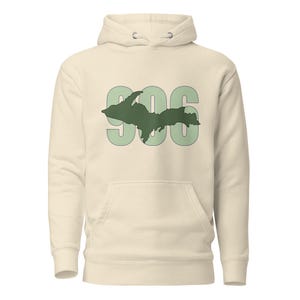 Puede incluir: Una sudadera con capucha de color crema que presenta un gráfico grande de color verde claro con los números 906 y una silueta verde oscuro de la península superior de Michigan superpuesta en el centro. Incluye un bolsillo tipo canguro.