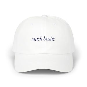 May include: A white baseball cap with a curved brim. The text "stack bestie" is embroidered in navy blue on the front of the cap. The cap has a rounded crown and a button on top.