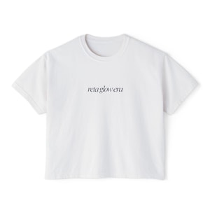 May include: A white, short-sleeved t-shirt with a crew neck. The text "reta glow era" is printed in a delicate, cursive font across the chest. The shirt is made of a soft, opaque fabric.
