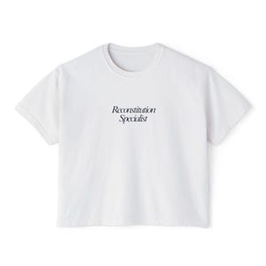 May include: A white, short-sleeved t-shirt with the text "Reconstitution Specialist" printed in black. The shirt has a crew neck and a relaxed fit, suitable for casual wear.