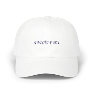 May include: White baseball cap with a curved brim. The cap features the text "reta glow era" in a dark blue, stylized font across the front. The hat is a classic six-panel design.
