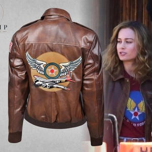 Può includere: Giacca bomber in pelle marrone dal design classico. Presenta una toppa rotonda sul retro con un emblema alato e un'illustrazione di aereo vintage. Include una toppa con la bandiera americana sulla manica superiore sinistra.