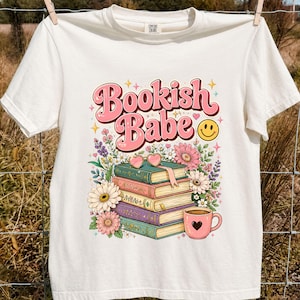 May include: An off-white short-sleeve t-shirt featuring a graphic print with the text Bookish Babe in pink letters. The design includes a stack of four books, a pink heart-shaped mug, colorful flowers, and a yellow smiley face icon.