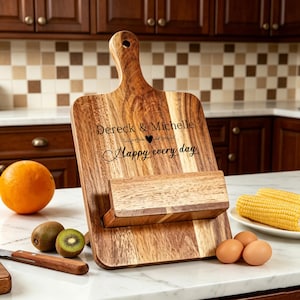 May include: Wooden cutting board with a handle and a built-in stand. The board is engraved with the names "Dereck & Michelle" and the phrase "Happy every day". The board is made of light and dark brown wood.