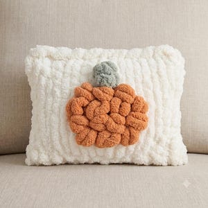 May include: A rectangular throw pillow made of white chunky yarn with a textured surface. Centered on the pillow is a pumpkin shape crafted from orange yarn loops, topped with a small stem made of sage green yarn loops.